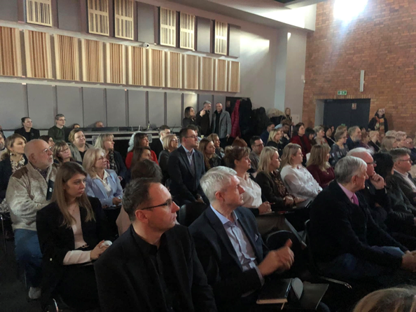 Na zdjęciu osoby siedzące w rzędach na krzesłach wsłuchujący się w przemówienie prowadzących